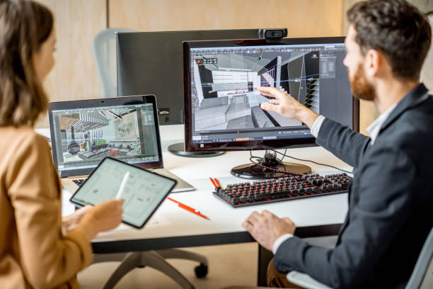 Two building designers for Spearfish Building Supply collaborate at a workstation, reviewing a 3D architectural model on a desktop monitor, with supporting visuals on a laptop and tablet, in a modern office setting.