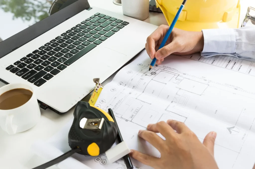 Close-up of a contractor reviewing architectural blueprints and making notes with a pencil beside a laptop, tape measure, and coffee cup—representing construction estimate services offered by Spearfish Building Supply.