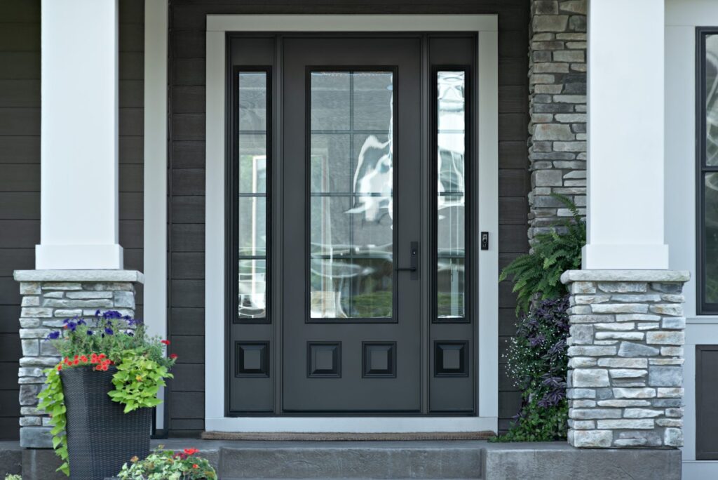 Front entry of a home featuring a sleek black exterior door with glass panels and matching sidelights, framed by white trim, stone columns, and vibrant potted plants—highlighting modern exterior door options with Spearfish Building Supply.