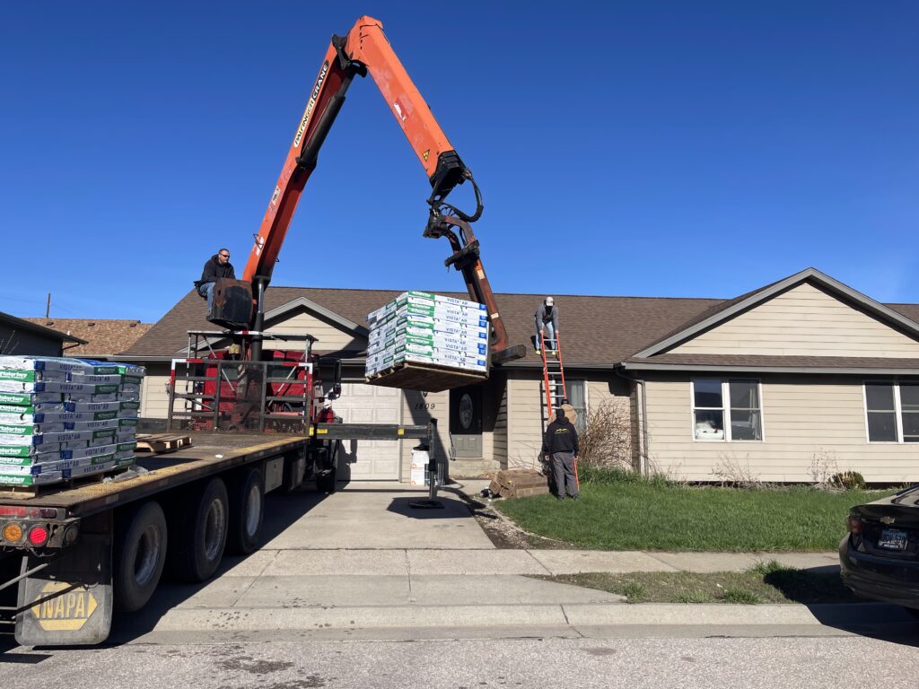 Construction crew using a crane truck to deliver and lift roofing materials onto a residential roof during a home improvement project with building materials delivery services with Spearfish Building Supply in Spearfish, SD.