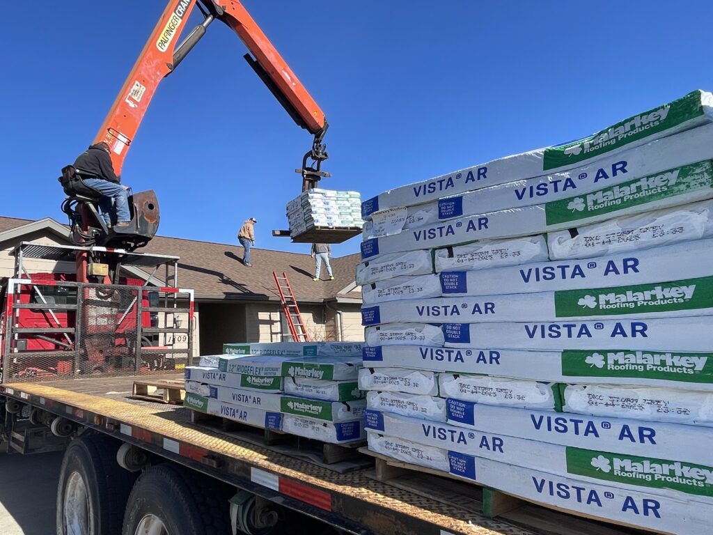 A Spearfish Building Supply forklift operator delivers stacked building materials at a residential construction site, with a partially framed house and clear blue sky in the background.