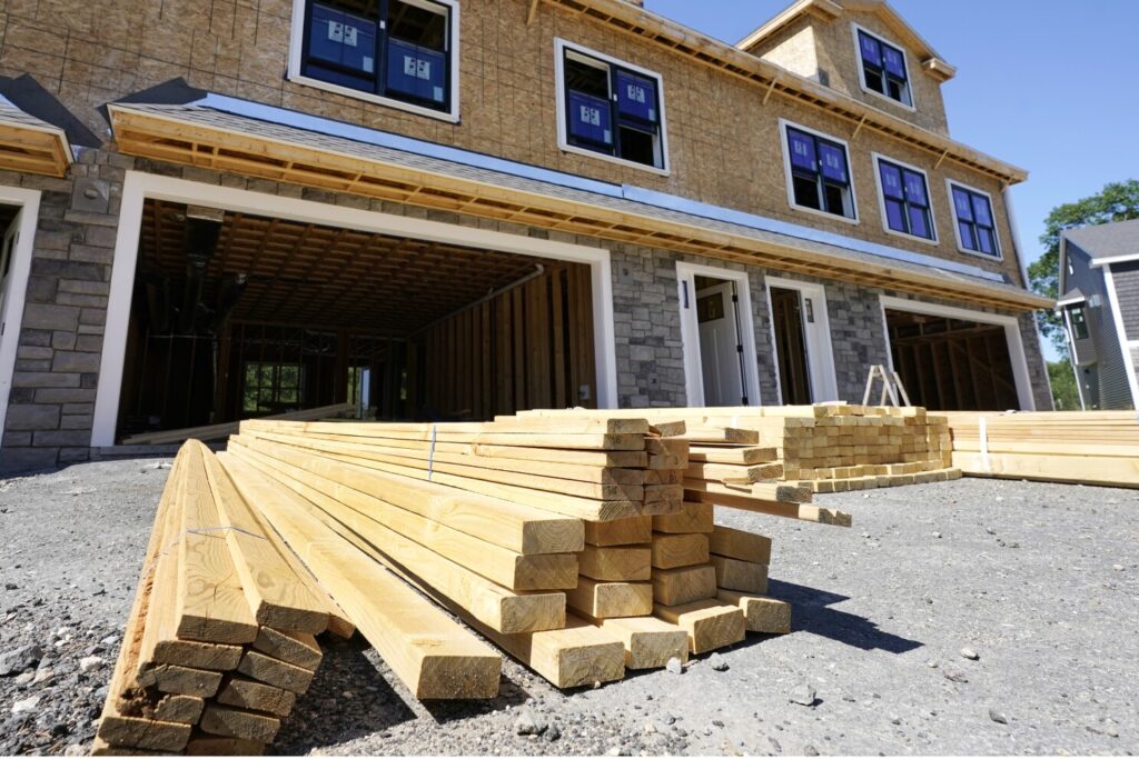 Stacks of lumber delivered in front of a multi-car garage at a residential construction site, with a partially framed house and exposed sheathing, illustrating new construction estimates and material supply services.