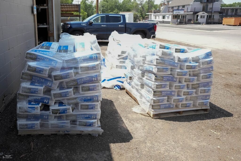 Pallets of Sakrete 60 lb Mortar Mix Type N bags wrapped in plastic and stacked outside a building supply store with a pickup truck parked behind.